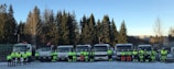 A group of people wearing reflective safety gear stand in front of a row of large trucks. The scene is set outdoors with coniferous trees in the background. The individuals are evenly spaced and seem to be part of a work crew, possibly for construction or transportation.