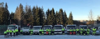 A group of people wearing reflective safety gear stand in front of a row of large trucks. The scene is set outdoors with coniferous trees in the background. The individuals are evenly spaced and seem to be part of a work crew, possibly for construction or transportation.