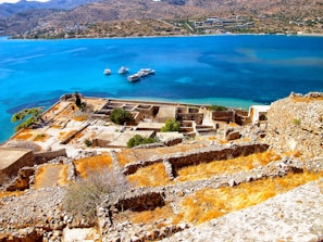 Overlooking ancient ruins with rocky stone walls and grassy patches set against a stunning deep blue sea. In the background, several boats float on the water while a hilly terrain lines the horizon.