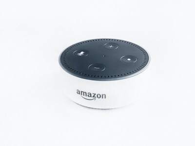 A small, cylindrical electronic device with a black top and white body, featuring several buttons for control functions. The top surface has a microphone button, volume control buttons, and a circular light indicator. The brand logo is displayed on the front.