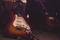 A sunburst electric guitar with a white pickguard is leaning against a vintage amplifier. The setting is dimly lit, suggesting a stage or rehearsal space, with another guitar partially visible in the background. The focus is on the guitar in the foreground, emphasizing its curves and finish.