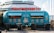 A retro-style turquoise food truck is parked in front of a building. The truck has vibrant red illuminated letters on top spelling out 'BRATYA GRILL' in Cyrillic. The sides of the truck are adorned with illustrations and names of different burger options, including Lucky, Killer, Chicken, Asia, Mona Lisa, Vegan, and Cowboy. Three wooden tables with metal barrels as bases are placed in front of the truck.