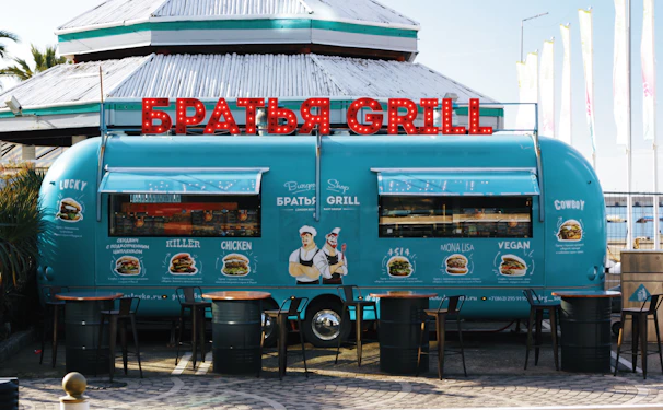 A retro-style turquoise food truck is parked in front of a building. The truck has vibrant red illuminated letters on top spelling out 'BRATYA GRILL' in Cyrillic. The sides of the truck are adorned with illustrations and names of different burger options, including Lucky, Killer, Chicken, Asia, Mona Lisa, Vegan, and Cowboy. Three wooden tables with metal barrels as bases are placed in front of the truck.