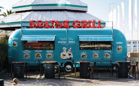 A retro-style turquoise food truck is parked in front of a building. The truck has vibrant red illuminated letters on top spelling out 'BRATYA GRILL' in Cyrillic. The sides of the truck are adorned with illustrations and names of different burger options, including Lucky, Killer, Chicken, Asia, Mona Lisa, Vegan, and Cowboy. Three wooden tables with metal barrels as bases are placed in front of the truck.