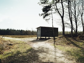 A rustic wooden shelter stands in a natural landscape, surrounded by sparse trees and a dirt path. The sun casts long shadows and lights up the open field behind the structure. The scene is tranquil with a touch of wilderness.