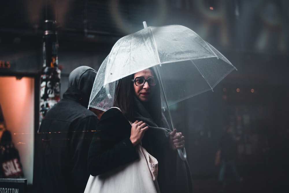 selective focus photography of woman carrying white bag while holding clear umbrella near person wearing hoodie outdoor