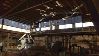 An installation of white bird sculptures is suspended from the ceiling of an industrial space. The setting features a high ceiling and large windows with gridded panes that allow natural light to enter. The interior is marked by metal beams and rustic brick walls, suggesting a workshop or factory environment.