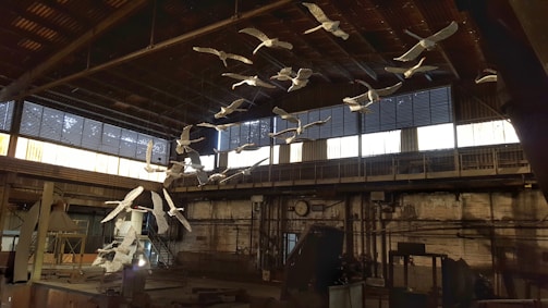 An installation of white bird sculptures is suspended from the ceiling of an industrial space. The setting features a high ceiling and large windows with gridded panes that allow natural light to enter. The interior is marked by metal beams and rustic brick walls, suggesting a workshop or factory environment.