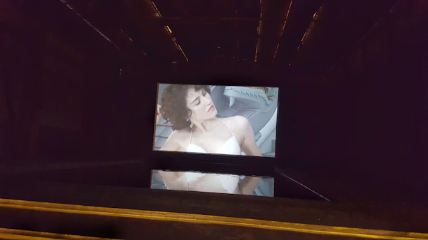 A dimly lit room features a large screen projecting the image of a person with curly hair and wearing a white top. The image appears to be from a film or video, with the person seated in what looks like a futuristic or modern chair. The screen's reflection is visible on a glossy surface below.