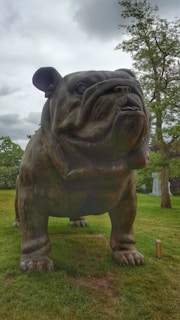 A large bronze sculpture of a bulldog stands prominently on a grassy area, surrounded by trees. The sky is overcast, giving the scene a slightly somber ambiance. The sculpture is detailed, capturing the sturdy build and distinctive features of a bulldog.