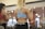 Two women are exercising in a gym, focusing on weightlifting with dumbbells. They are both wearing athletic clothing; one in a blue top and the other in a red top. Their reflections are visible in the mirror behind them, and the background is softly blurred with gym equipment visible.