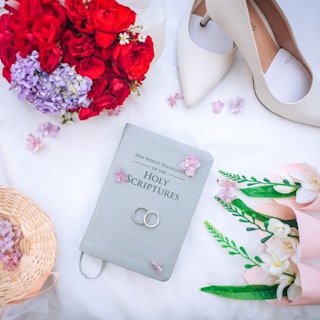 A beautifully composed arrangement consisting of a Bible labeled 'New World Translation of the Holy Scriptures', wedding rings placed on the Bible, white high-heeled shoes, red and purple flowers, a wicker basket, and a bouquet of white flowers wrapped in pink. The items are set on a white surface scattered with small pink flower petals.