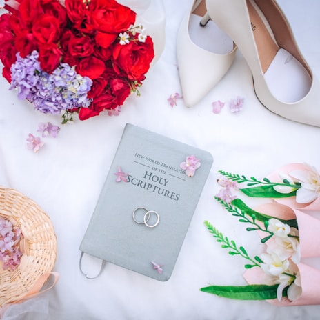 A beautifully composed arrangement consisting of a Bible labeled 'New World Translation of the Holy Scriptures', wedding rings placed on the Bible, white high-heeled shoes, red and purple flowers, a wicker basket, and a bouquet of white flowers wrapped in pink. The items are set on a white surface scattered with small pink flower petals.