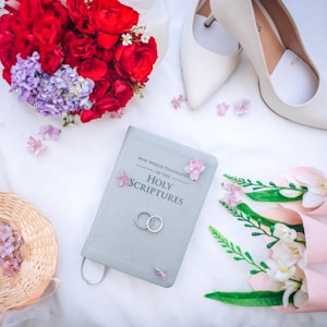 A beautifully composed arrangement consisting of a Bible labeled 'New World Translation of the Holy Scriptures', wedding rings placed on the Bible, white high-heeled shoes, red and purple flowers, a wicker basket, and a bouquet of white flowers wrapped in pink. The items are set on a white surface scattered with small pink flower petals.