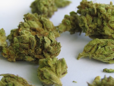 A close-up view of several cannabis buds, showing their intricate, leafy structures with hues of green and orange. The surface texture appears frosty and glandular due to the presence of trichomes.