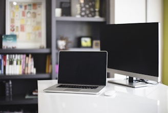 A modern workspace featuring a laptop and monitor on a white desk. The background includes a bookshelf filled with books and decorative items.