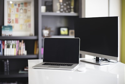 A modern workspace featuring a laptop and monitor on a white desk. The background includes a bookshelf filled with books and decorative items.