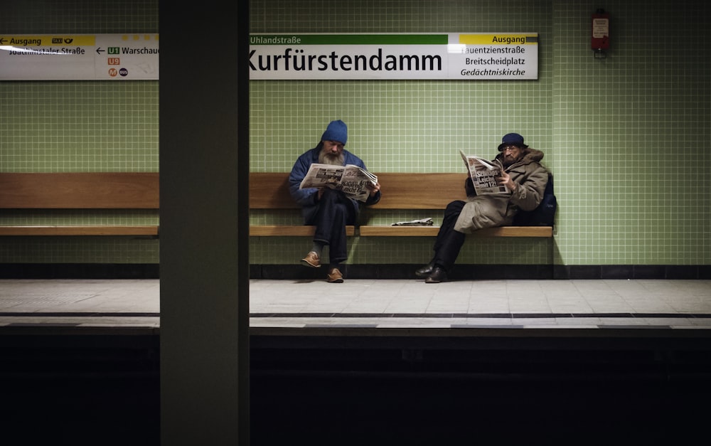 zwei Männer sitzen auf brauner Holzbank und lesen Zeitschriften