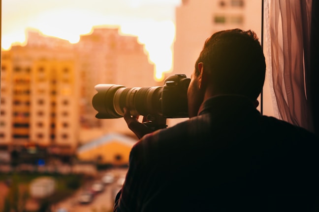 A person is holding a large camera with a telephoto lens, looking out of a window at a cityscape during sunset. The buildings and sky are illuminated with warm golden and orange tones, creating a backlit silhouette of the individual.