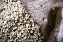 A burlap sack partially filled with raw, unroasted coffee beans. The coffee beans are light greenish-beige in color and are spilling out of the sack onto a textured surface.
