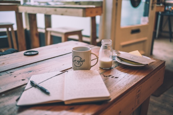 A wooden table holds an open notebook with a pen resting on it. Nearby, a ceramic mug with writing is positioned beside a small glass milk bottle. In the background, a plate with a napkin and cutlery sits near a bright window, creating a warm and inviting atmosphere.
