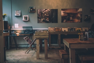 A cozy interior features a wooden floor and rustic decor. Vinyl records are displayed on a wooden shelf, with a record player and a small table nearby. Several framed pictures hang on the wall. A small vase with flowers sits on a table, adding a touch of color to the warm, dimly lit atmosphere.