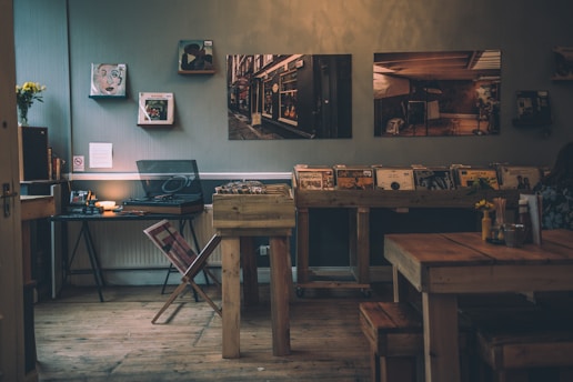 A cozy interior features a wooden floor and rustic decor. Vinyl records are displayed on a wooden shelf, with a record player and a small table nearby. Several framed pictures hang on the wall. A small vase with flowers sits on a table, adding a touch of color to the warm, dimly lit atmosphere.