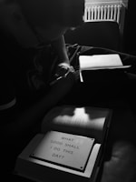 A person is writing in a notebook while sitting in a dimly lit room. The focus is on an open book with a prominent sign that reads 'What good shall I do this day?', highlighting a contemplative atmosphere. The overall setting is subdued, with shadows and soft light creating a reflective mood.