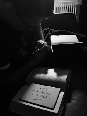 A person is writing in a notebook while sitting in a dimly lit room. The focus is on an open book with a prominent sign that reads 'What good shall I do this day?', highlighting a contemplative atmosphere. The overall setting is subdued, with shadows and soft light creating a reflective mood.