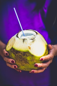 A person is holding a fresh green coconut with a straw inserted into it. The person's nails are painted a deep red color, and they are wearing a purple garment.