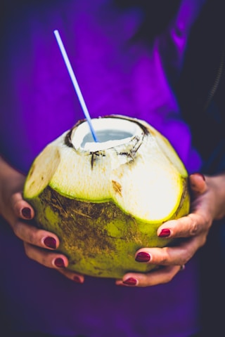 A person is holding a fresh green coconut with a straw inserted into it. The person's nails are painted a deep red color, and they are wearing a purple garment.