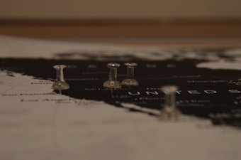 A close-up view of a map featuring the United States with several transparent push pins marking specific locations. The map is predominantly black and white, with the push pins adding a subtle three-dimensional element.