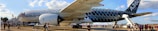 A large Airbus A350 aircraft is positioned on an airport tarmac with visible landing gear and engine details. The fuselage is decorated with a distinctive pattern, and it is surrounded by people, possibly visitors or aviation enthusiasts. A set of stairs leads up to the cabin doors, and the scene takes place in daylight under a partly cloudy sky.