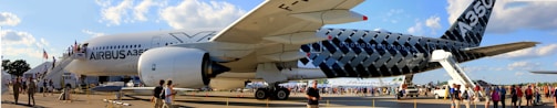 A large Airbus A350 aircraft is positioned on an airport tarmac with visible landing gear and engine details. The fuselage is decorated with a distinctive pattern, and it is surrounded by people, possibly visitors or aviation enthusiasts. A set of stairs leads up to the cabin doors, and the scene takes place in daylight under a partly cloudy sky.