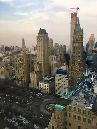 A cityscape features a cluster of tall, historic buildings with ornate architectural details and a construction crane towering above them. Below, a bustling street filled with vehicles, including yellow taxis, is visible. The surrounding area includes a lightly snow-dusted park with bare trees, indicating a cold season.