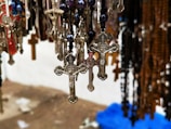 Hanging metal crucifixes and rosaries with detailed designs are shown in a close-up view. The background appears to be blurry, showcasing even more rosaries and crucifixes of various colors and materials.