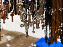 Hanging metal crucifixes and rosaries with detailed designs are shown in a close-up view. The background appears to be blurry, showcasing even more rosaries and crucifixes of various colors and materials.