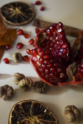 A close-up view of pomegranate seeds and dried fruits elegantly arranged on a light surface. The vibrant red seeds contrast with the muted tones of the dried citrus slices and other textured botanicals.