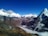 A stunning Himalayan mountain landscape with snow-capped peaks under a clear blue sky. The foreground shows rugged, rocky terrain, leading into deep valleys covered by layers of low clouds. The majestic mountains dominate the scene with their white, glistening snow and steep, dark slopes.