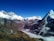 A stunning Himalayan mountain landscape with snow-capped peaks under a clear blue sky. The foreground shows rugged, rocky terrain, leading into deep valleys covered by layers of low clouds. The majestic mountains dominate the scene with their white, glistening snow and steep, dark slopes.