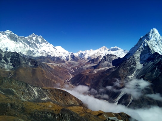 A stunning Himalayan mountain landscape with snow-capped peaks under a clear blue sky. The foreground shows rugged, rocky terrain, leading into deep valleys covered by layers of low clouds. The majestic mountains dominate the scene with their white, glistening snow and steep, dark slopes.