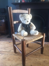A fluffy white teddy bear with a dark bow sits on a woven wooden chair in a dimly lit room. The chair has a simple design with a tall back and a woven seat. Behind the chair, a dark fireplace and some pieces of furniture are visible.