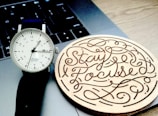 A wristwatch with a minimalistic design, featuring a white face and black numbers, sits on a laptop keyboard next to a wooden coaster with the words 'Stay Focused' intricately carved into it.