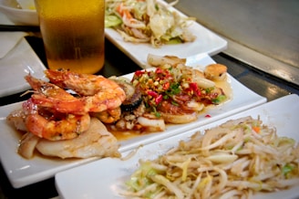 A serving of various types of food set on white rectangular plates. There are cooked prawns placed atop white fish fillets, a mixed stir-fry of meat, vegetables, and peppers with red chili, and a plate of stir-fried bean sprouts with shredded vegetables. A cold glass of beer is partially visible, along with more food items in the background.