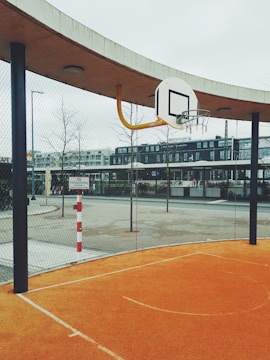 A basketball hoop is set against an orange outdoor court with a metal fence surrounding the area. Several buildings are visible in the background, and the environment appears urban with overcast skies.