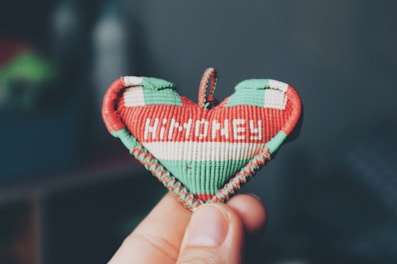 A hand is holding a small, heart-shaped object made of woven material. The object has a red, white, and green color scheme and the word 'HUNGARY' is embroidered in white on it. The background is blurred and appears to be indoors.