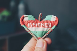 A hand is holding a small, heart-shaped object made of woven material. The object has a red, white, and green color scheme and the word 'HUNGARY' is embroidered in white on it. The background is blurred and appears to be indoors.