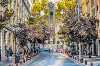 A bustling urban street scene featuring a tall, industrial-looking elevator structure at the center, surrounded by trees and historic buildings. People are walking along the sidewalks, and several vehicles are parked or moving on the street. The sun is casting soft light, creating a warm atmosphere.