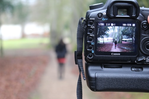 A digital camera is positioned in the foreground with the viewfinder screen displaying an image of a person standing on a path in a park. The background outside the camera display is blurred, featuring the same path and surrounding greenery.