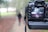 A digital camera is positioned in the foreground with the viewfinder screen displaying an image of a person standing on a path in a park. The background outside the camera display is blurred, featuring the same path and surrounding greenery.